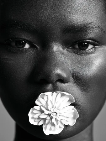 Close-up portrait of an African American woman with dark skin, a flower in her mouth, captured in black and white with a soft gray background.
