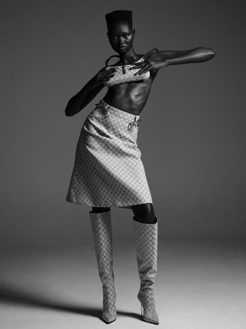 Monochrome studio shot of an adult female fashion model posing in a patterned skirt, tube top, and knee-high boots while holding a purse.