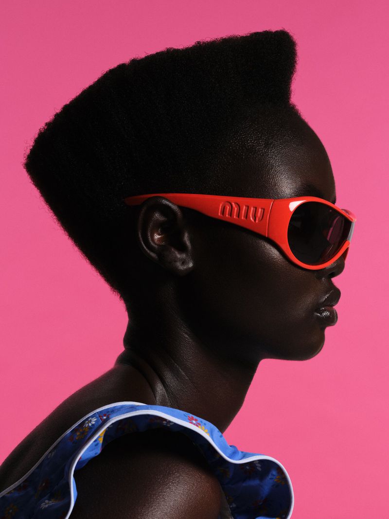 Close-up of an adult African-descent woman with a high-top fade haircut wearing orange sunglasses against a bright pink background.