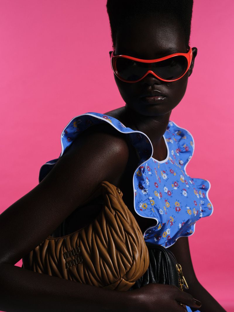 A woman with dark skin poses in a light blue, floral-patterned dress and orange sunglasses, holding a caramel-colored purse against a pink backdrop.