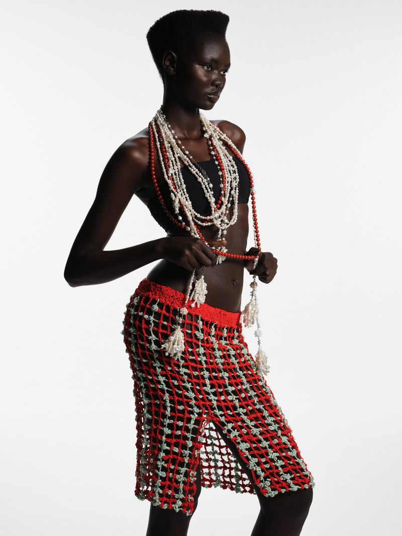 A dark-skinned woman stands in a studio wearing a crochet skirt and multiple beaded necklaces against a bright, neutral background.