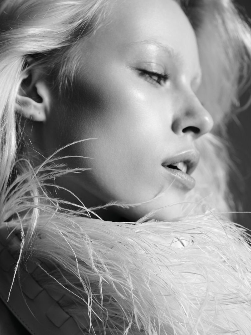 Black and white portrait of a young woman with pale skin, light eyebrows and light blonde hair wearing white feathers around her shoulders.