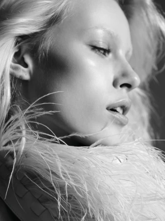Black and white portrait of a young woman with pale skin, light eyebrows and light blonde hair wearing white feathers around her shoulders.