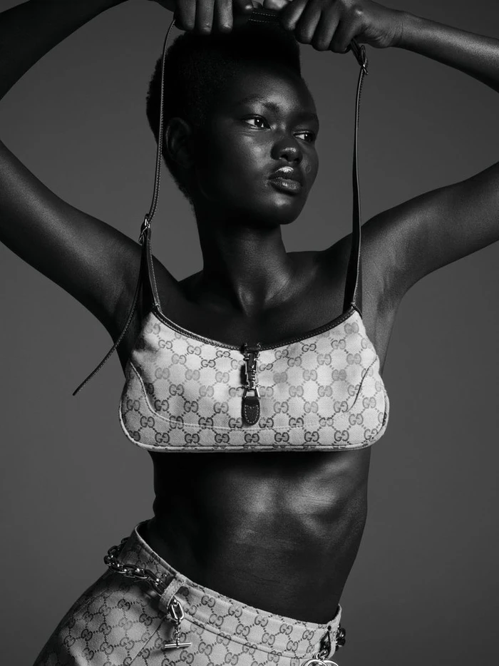Black and white fashion portrait of a model holding a patterned shoulder bag overhead against a dark studio background