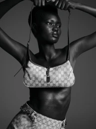 Black and white fashion portrait of a model holding a patterned shoulder bag overhead against a dark studio background