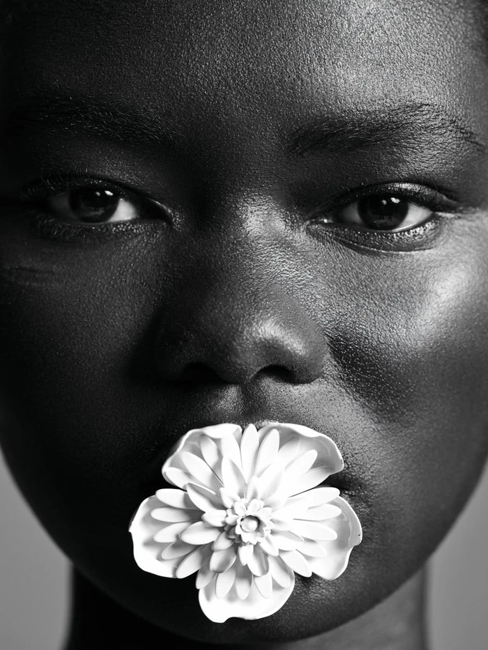 Extreme close up portrait of a Black woman holding a white ceramic flower in her mouth.