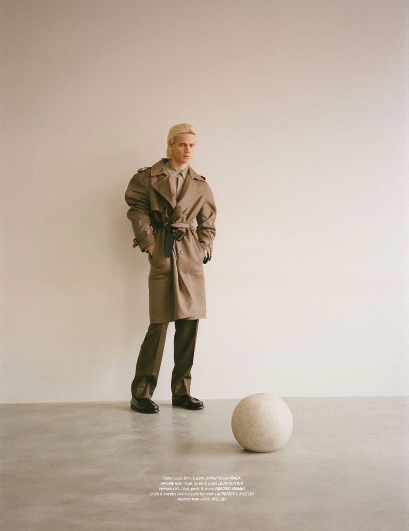 Fashion editorial shot of a blonde male model in a tan trench coat standing indoors with a neutral background and a round stone prop.