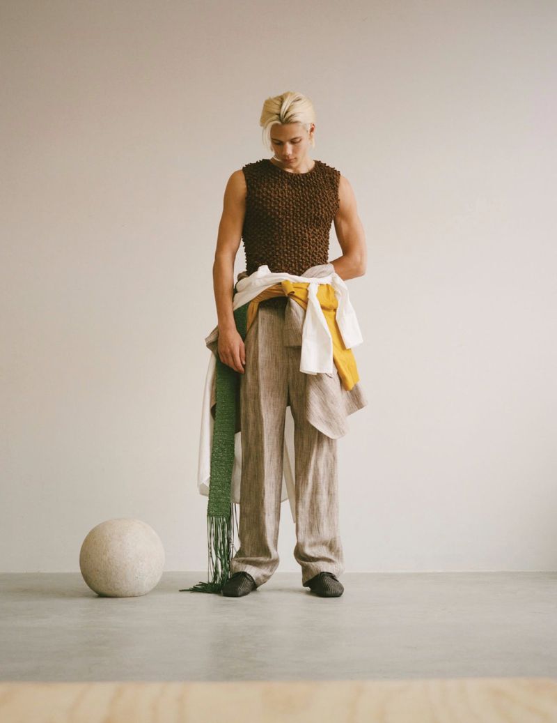 Blond male model wearing a brown knit tank top and baggy linen pants stands in a photo studio with neutral color tones.