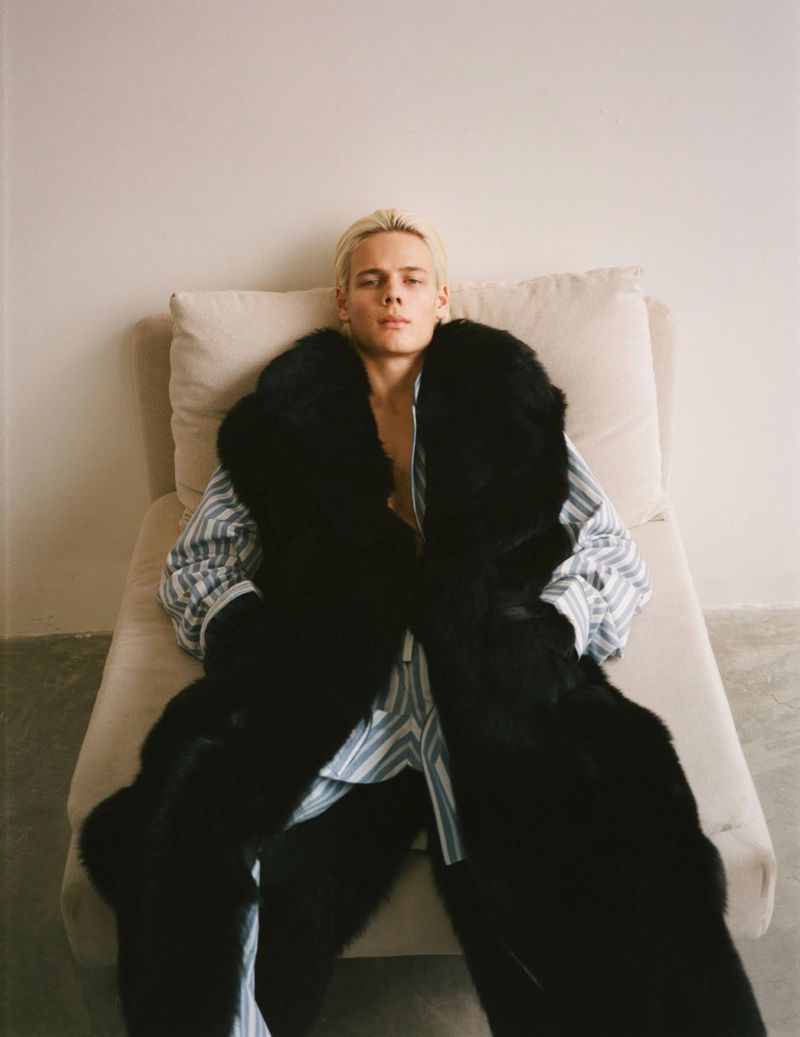Fashion portrait of a blonde adult male in pajamas and a dark fur coat reclining on a light colored lounge indoors with a neutral background.