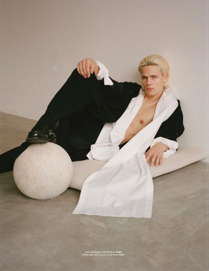 Male fashion model with blond hair poses in a black suit and white shirt, reclining on a cushion with a stone sphere in studio setting.