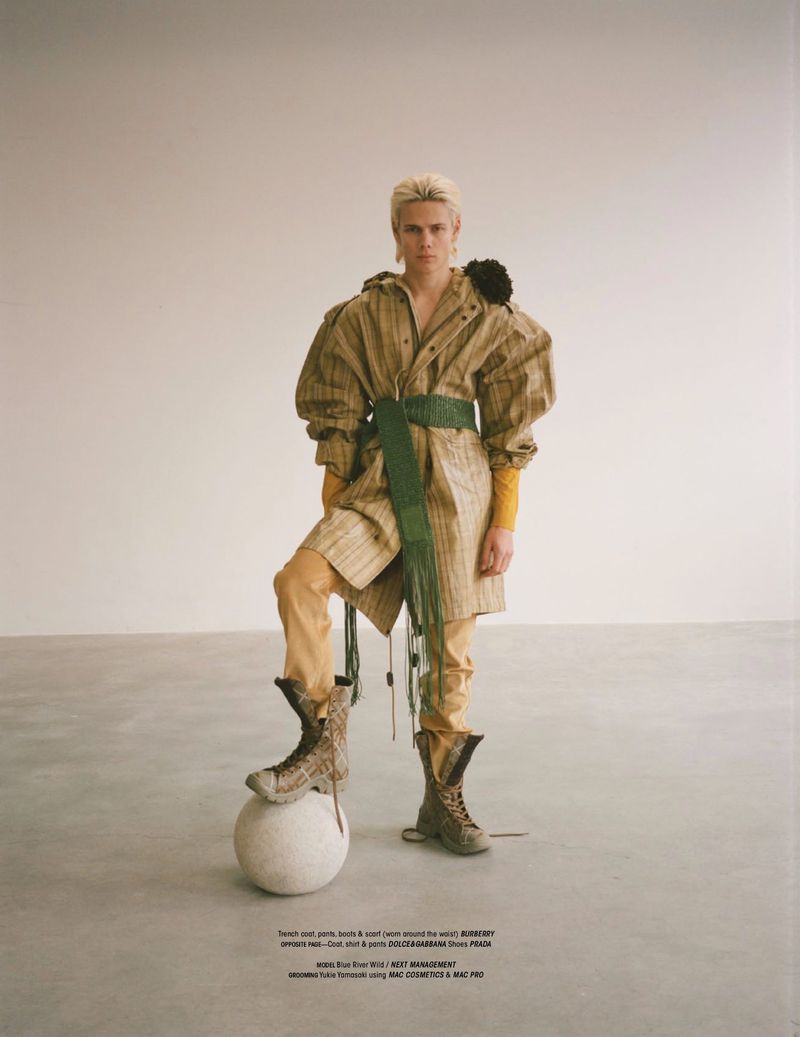 Fashion editorial shot of a non-binary person in an over-sized tan trench coat and combat boots posing with a light-gray sphere indoors.