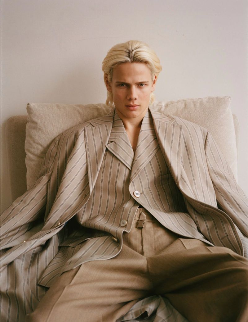 Portrait of a fair-skinned young adult male with blonde hair wearing a striped tan suit jacket and pants, and sitting on a cushion.