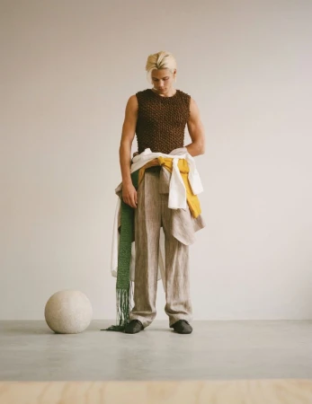 Blonde male model wearing a brown knit top and beige trousers standing against a seamless grey backdrop with a sphere prop
