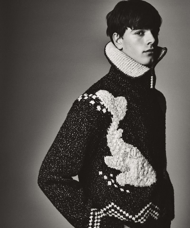 Monochromatic portrait of a young man wearing a patterned wool sweater with a high collar, eyes forward, in a studio setting.