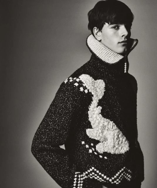 Monochromatic portrait of a young man wearing a patterned wool sweater with a high collar, eyes forward, in a studio setting.