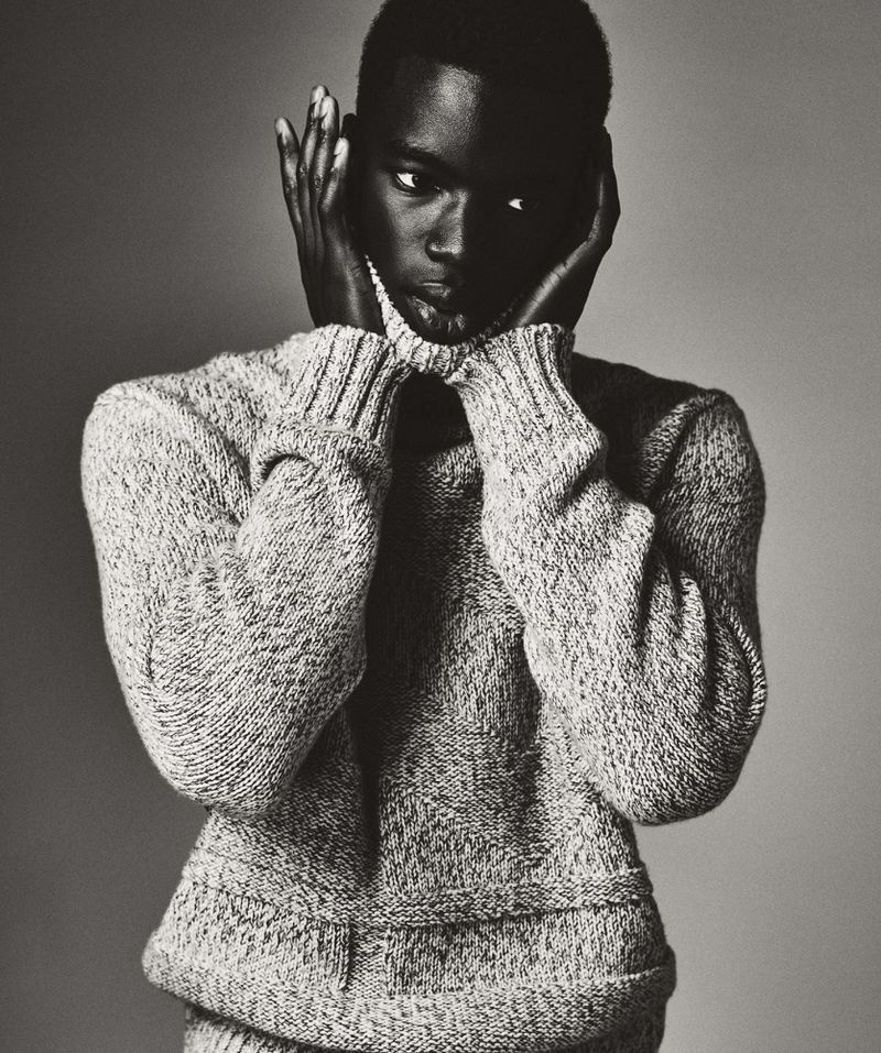Portrait of a dark-skinned male model wearing a gray turtleneck sweater, posing with hands near face, against a neutral gray backdrop.