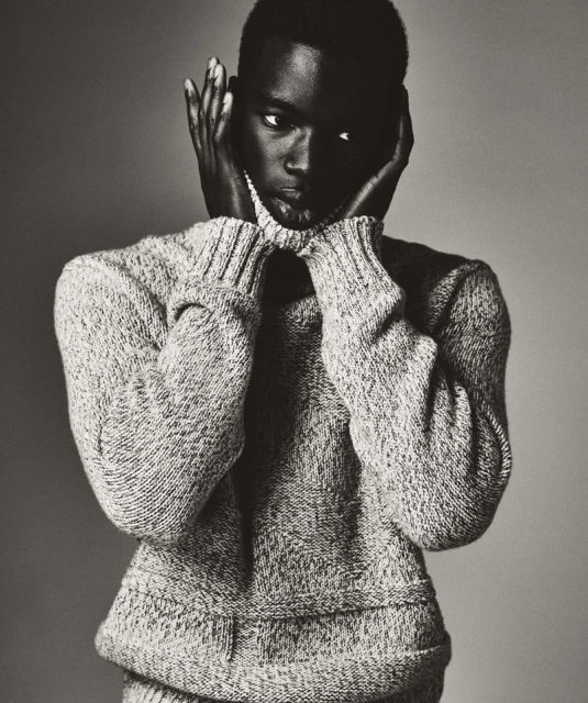 Portrait of a dark-skinned male model wearing a gray turtleneck sweater, posing with hands near face, against a neutral gray backdrop.