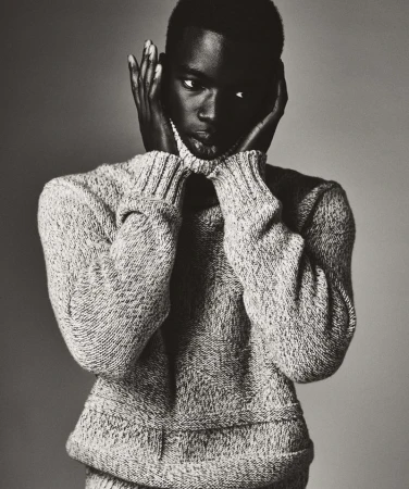 Portrait of a dark-skinned male model wearing a gray turtleneck sweater, posing with hands near face, against a neutral gray backdrop.