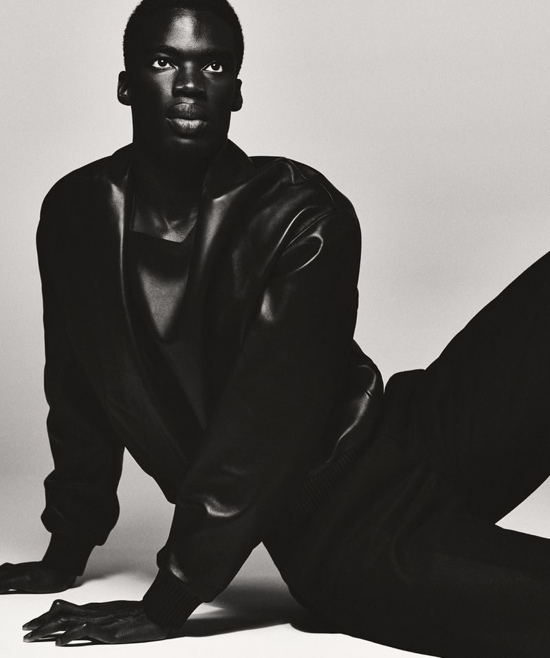 Portrait of a Black male model in a black leather jacket and black pants, kneeling against a light gray backdrop in a studio setting.