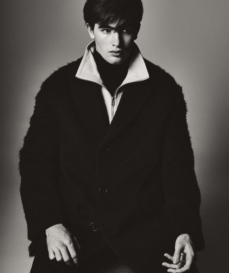 Portrait of a young adult male wearing a dark, textured coat over a turtleneck sweater in a studio with neutral background.