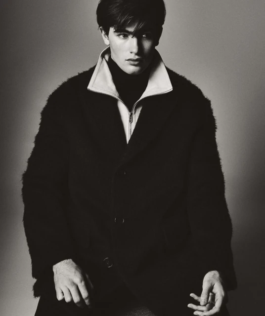 Portrait of a young adult male wearing a dark, textured coat over a turtleneck sweater in a studio with neutral background.