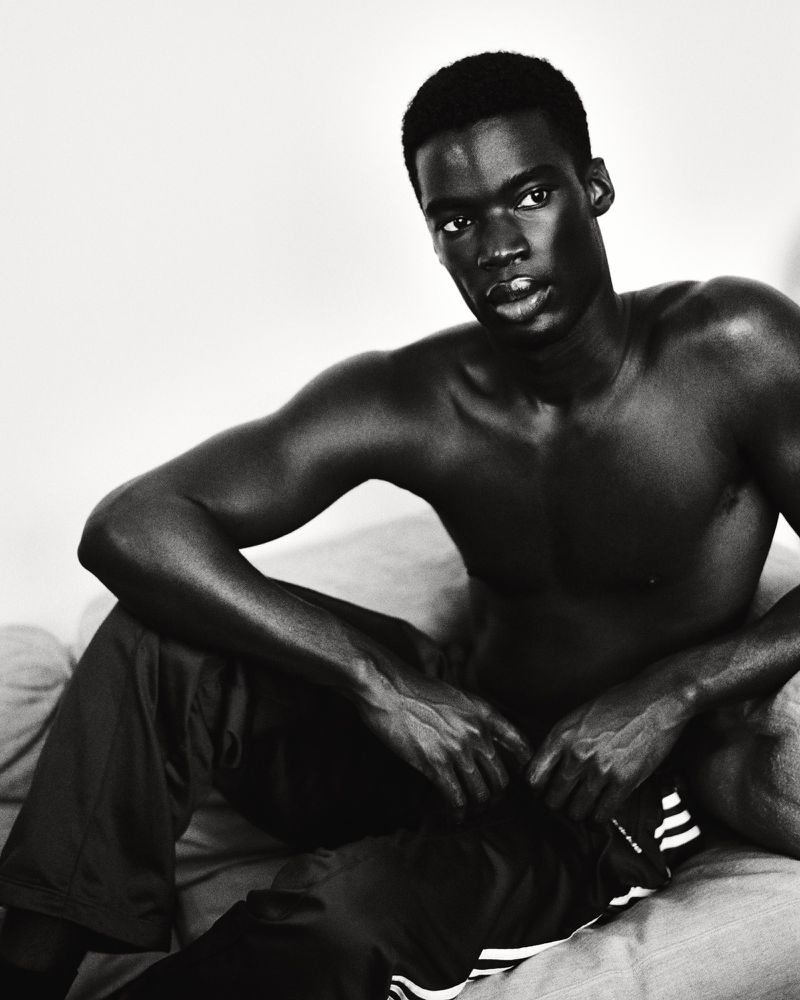 Monochrome portrait of bare-chested Black man seated, wearing Adidas pants, showcasing a serious expression in indoor setting.
