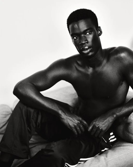Monochrome portrait of bare-chested Black man seated, wearing Adidas pants, showcasing a serious expression in indoor setting.