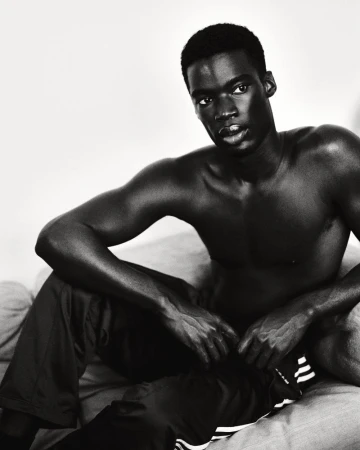 Monochrome portrait of bare-chested Black man seated, wearing Adidas pants, showcasing a serious expression in indoor setting.