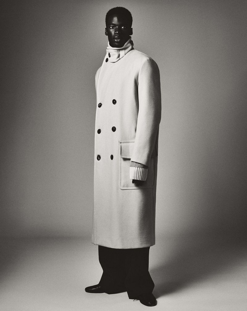 Full-length portrait of a Black male fashion model in a long double-breasted coat and black pants, posing in a studio setting with a gray background.