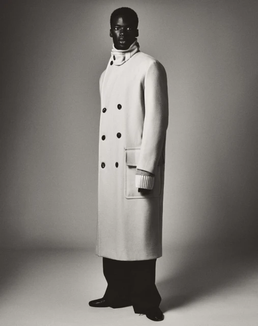 Full-length portrait of a Black male fashion model in a long double-breasted coat and black pants, posing in a studio setting with a gray background.