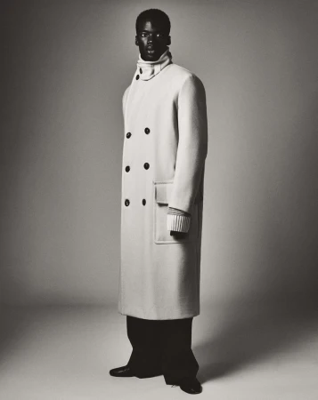 Full-length portrait of a Black male fashion model in a long double-breasted coat and black pants, posing in a studio setting with a gray background.