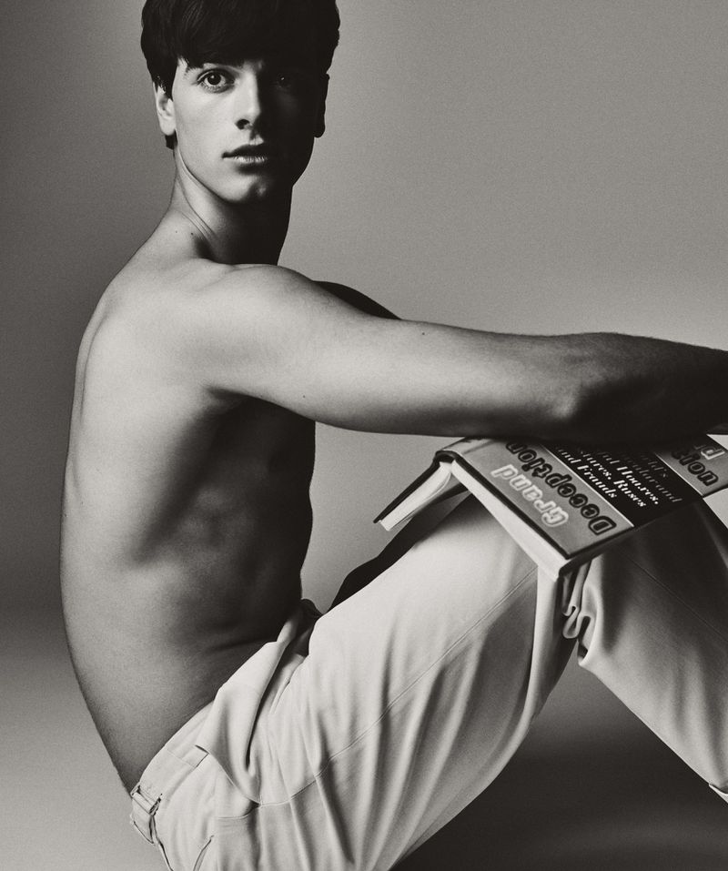 A shirtless young man with dark hair poses with a book in a studio setting with a neutral background in a black and white photograph.