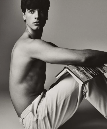 A shirtless young man with dark hair poses with a book in a studio setting with a neutral background in a black and white photograph.