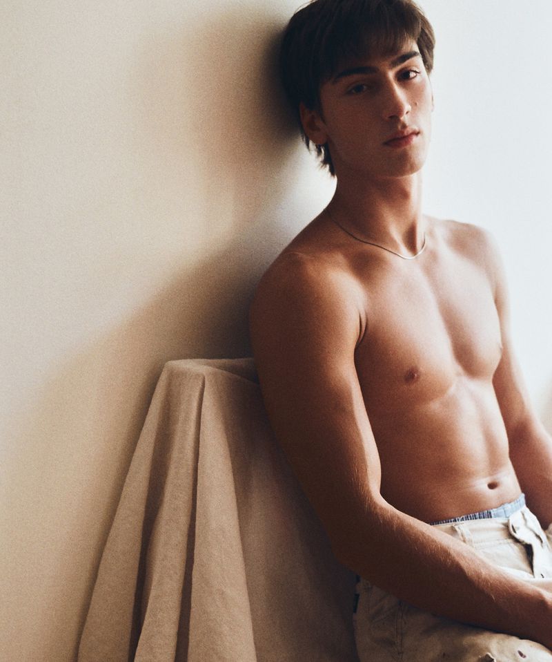 Shirtless young man leaning against a plain wall draped with fabric, photographed by Joe Brennan for HERO Magazine.