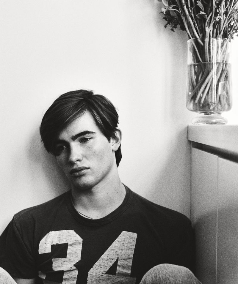 Monochrome portrait of a young man wearing a graphic t-shirt against a plain background with a glass vase of flowers.