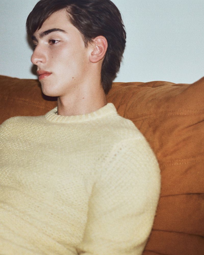 A young adult male with dark hair wearing a light yellow sweater is seated indoors, posing in a medium shot against a tan couch.