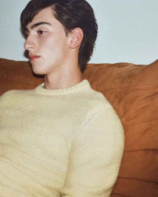 A young adult male with dark hair wearing a light yellow sweater is seated indoors, posing in a medium shot against a tan couch.
