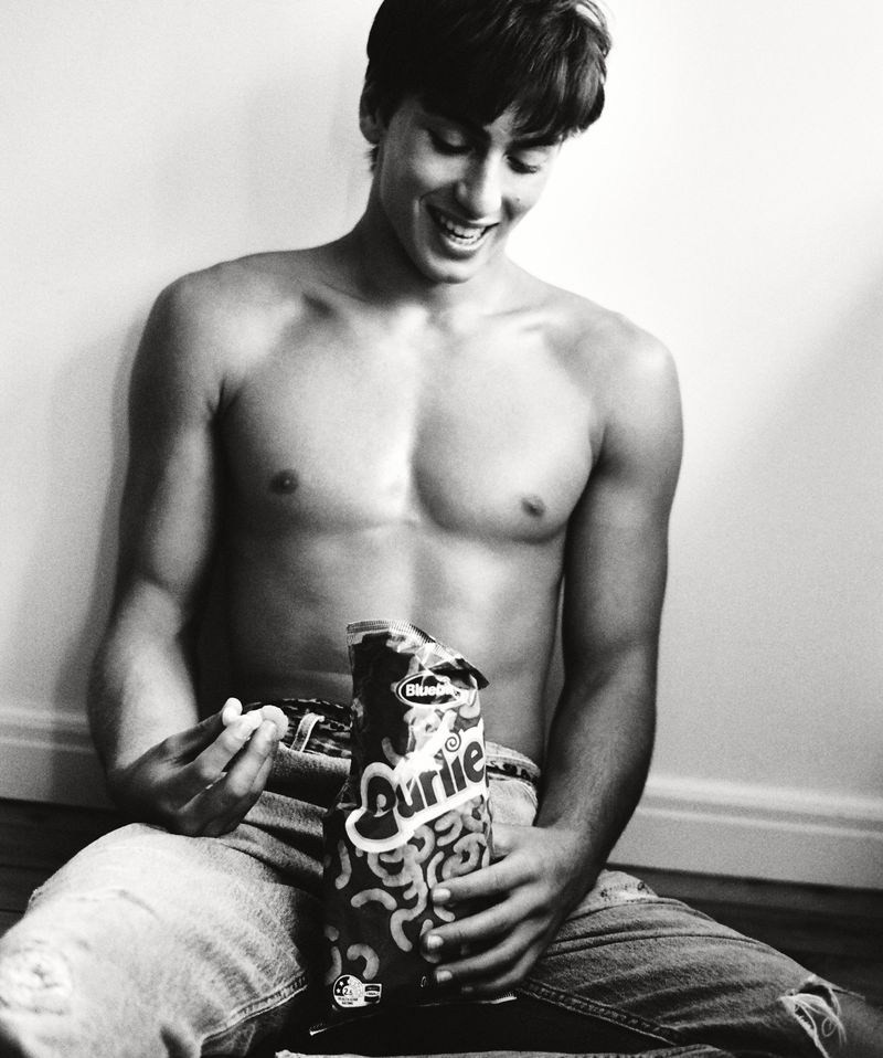 A topless young man wearing jeans sits against a wall and smiles as he holds a bag of snack food in a black-and-white photo.