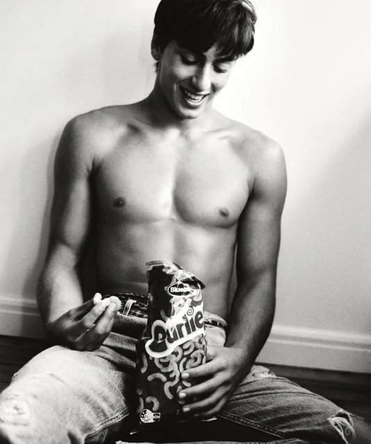 A topless young man wearing jeans sits against a wall and smiles as he holds a bag of snack food in a black-and-white photo.