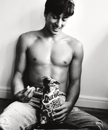 A topless young man wearing jeans sits against a wall and smiles as he holds a bag of snack food in a black-and-white photo.
