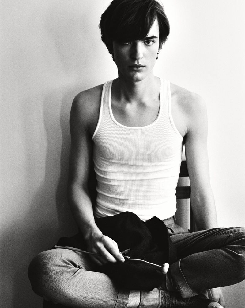 Portrait of a young adult male sitting on a chair with a black jacket in his hands, set against a simple white background.