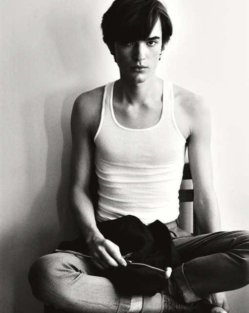 Portrait of a young adult male sitting on a chair with a black jacket in his hands, set against a simple white background.