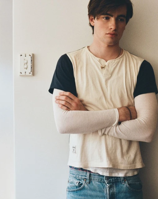 Portrait of a young adult male with arms crossed, wearing a light blue jeans and a cream and navy baseball tee, standing indoors.