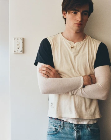 Portrait of a young adult male with arms crossed, wearing a light blue jeans and a cream and navy baseball tee, standing indoors.