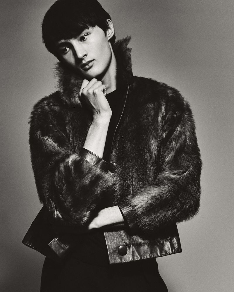 Portrait of Asian male model in black top and fur jacket, posing against grey studio background. Fashion, elegance, and sophistication.