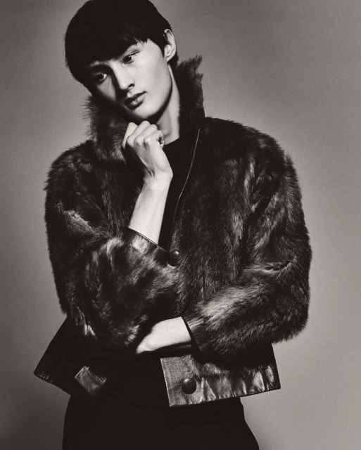 Portrait of Asian male model in black top and fur jacket, posing against grey studio background. Fashion, elegance, and sophistication.