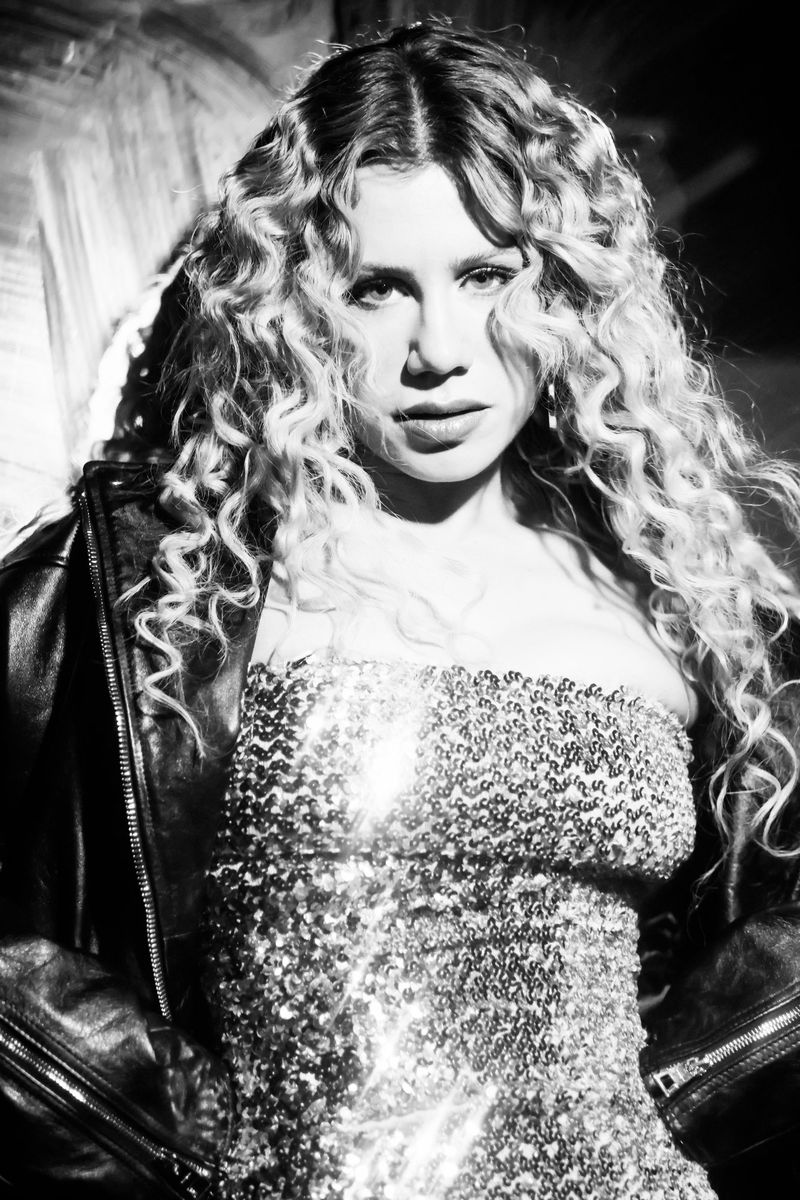 Black and white portrait of a woman with curly blonde hair wearing a sequined top and leather jacket against a textured background.
