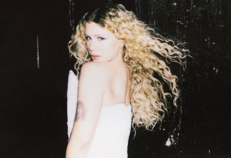 Blonde woman in a white dress gazes over her shoulder against a dark background with mottled texture and soft, diffused lighting.