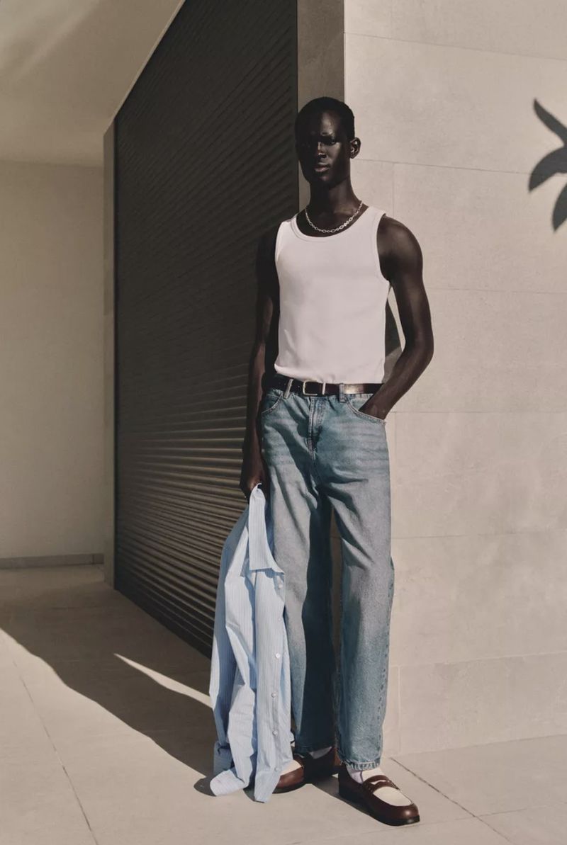 A dark-skinned male model is wearing a white tank top and jeans against a minimalist wall with shadows and holding a light-blue shirt.