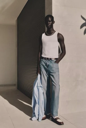A dark-skinned male model is wearing a white tank top and jeans against a minimalist wall with shadows and holding a light-blue shirt.